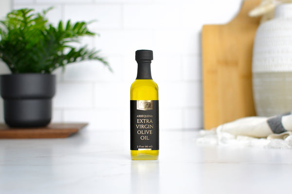 A bottle of Jocelyn & Co Arbequina Extra Virgin Olive Oil stands on a white countertop in a bright kitchen setting, with a blurred potted plant on the left and kitchen items on the right.