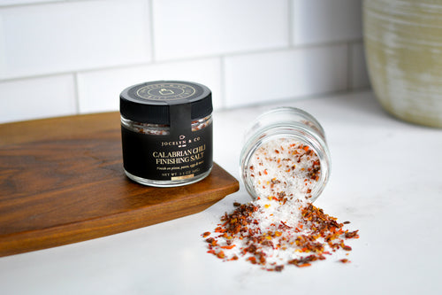 A small glass jar of Joccelyn & Co Calabrian Chili Finishing Salt jar stands on a wooden board with another tipped-over jar spilling white salt crystals mixed with red chili flakes onto a white countertop.