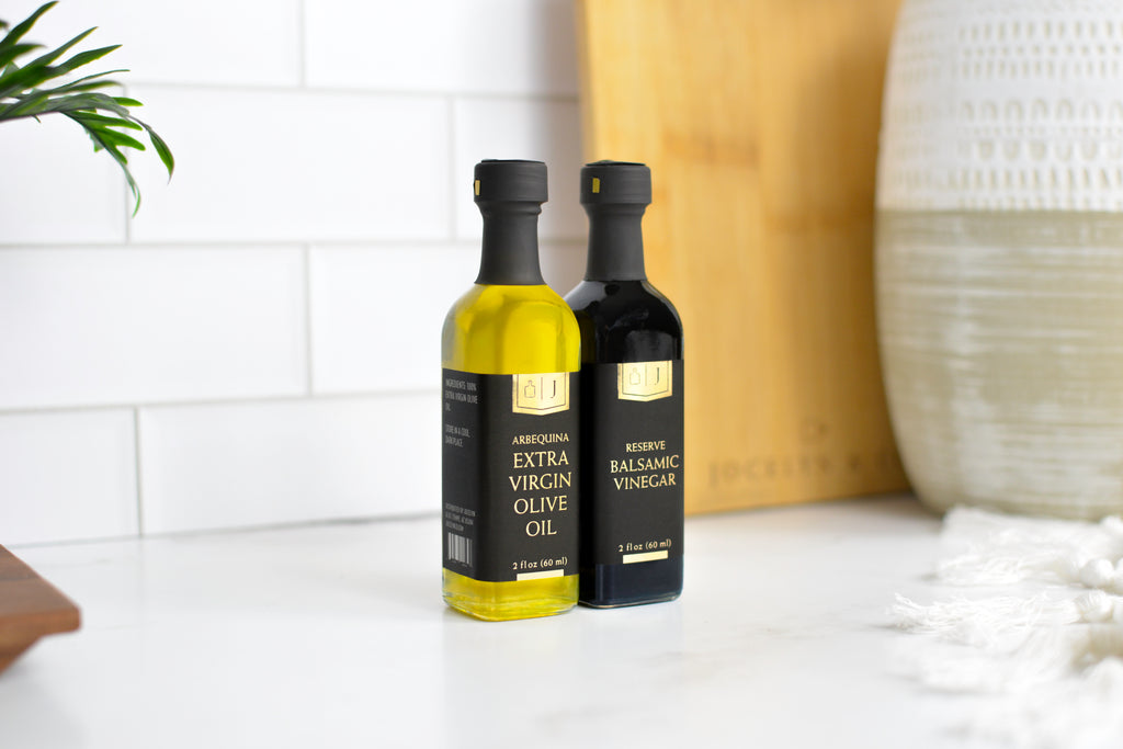 Two bottles of Jocelyn & co olive oil and balsamic vinegar standing on a white marble countertop against a white subway tile backsplash. The bottle on the left contains golden olive oil and is labeled 'Arbequina Extra Virgin Olive Oil', and the bottle on the right is dark brown/black and labeled 'Reserve Balsamic Vinegar'.