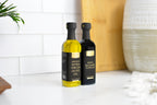 Two bottles of Jocelyn & co olive oil and balsamic vinegar standing on a white marble countertop against a white subway tile backsplash. The bottle on the left contains golden olive oil and is labeled 'Arbequina Extra Virgin Olive Oil', and the bottle on the right is dark brown/black and labeled 'Reserve Balsamic Vinegar'.