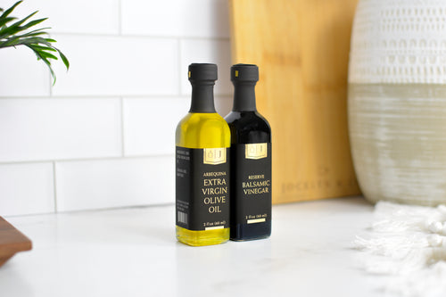 Two bottles of Jocelyn & co olive oil and balsamic vinegar standing on a white marble countertop against a white subway tile backsplash. The bottle on the left contains golden olive oil and is labeled 'Arbequina Extra Virgin Olive Oil', and the bottle on the right is dark brown/black and labeled 'Reserve Balsamic Vinegar'.