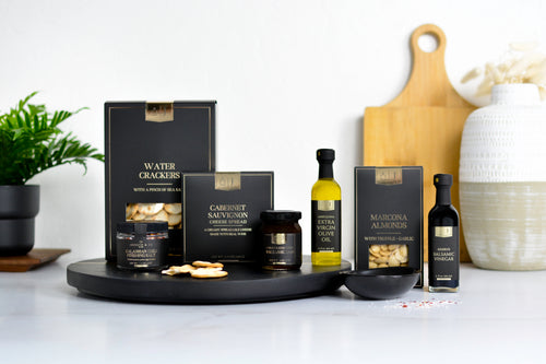 A gourmet food gift set from Jocelyn & Co, featuring their black & gold line with boxes and jars of water crackers, cabernet sauvignon cheese spread, garlic truffle marcona almonds, and olive oil, arranged on a black tray with a bowl and spices, set against a bright white background with wooden cutting board and a ceramic vase.