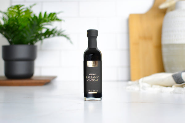 A bottle of Jocelyn & Co Premium Balsamic Vinegar standing on a white countertop in a bright kitchen setting with a blurred potted plant and wooden cutting board in the background.