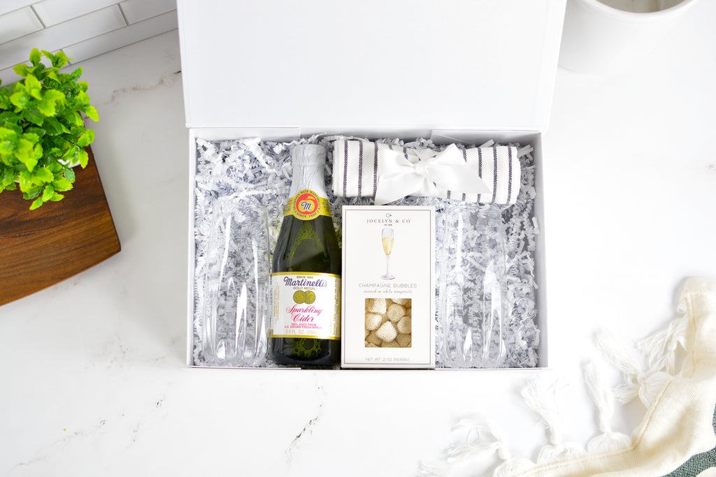 A curated gift box set on a white marble surface, featuring a bottle of sparkling wine, a boxed chocolate selection, and striped glassware nestled in white crinkle paper inside an open white box.