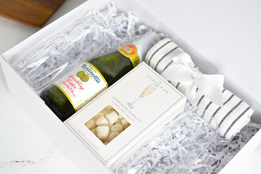 A luxury gift box set containing a bottle of Perigot champagne, a box of Truffauts cheese, and a striped white and green towel, all nestled in white shredded paper inside a white box.