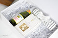 A luxury gift box set containing a bottle of Perigot champagne, a box of Truffauts cheese, and a striped white and green towel, all nestled in white shredded paper inside a white box.