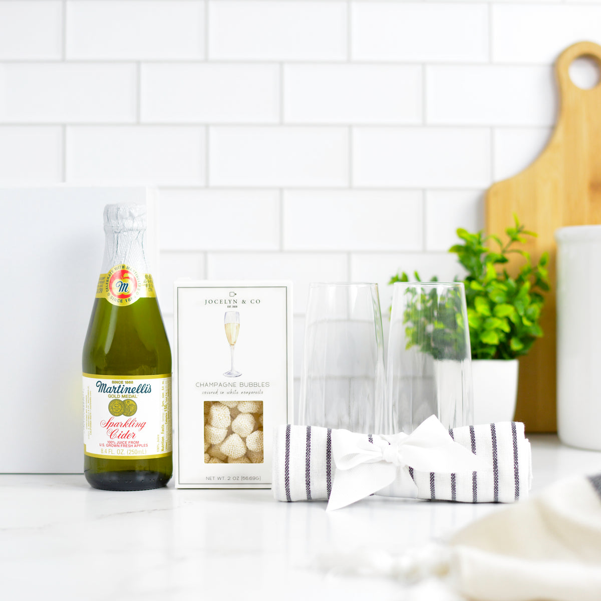 A champagne-themed gift set on a white countertop with a Martinelli sparkling cider bottle, a box of Fresh & Co champagne bubbles, two clear flutes, and a striped napkin tied with a bow, against a white subway tile background.