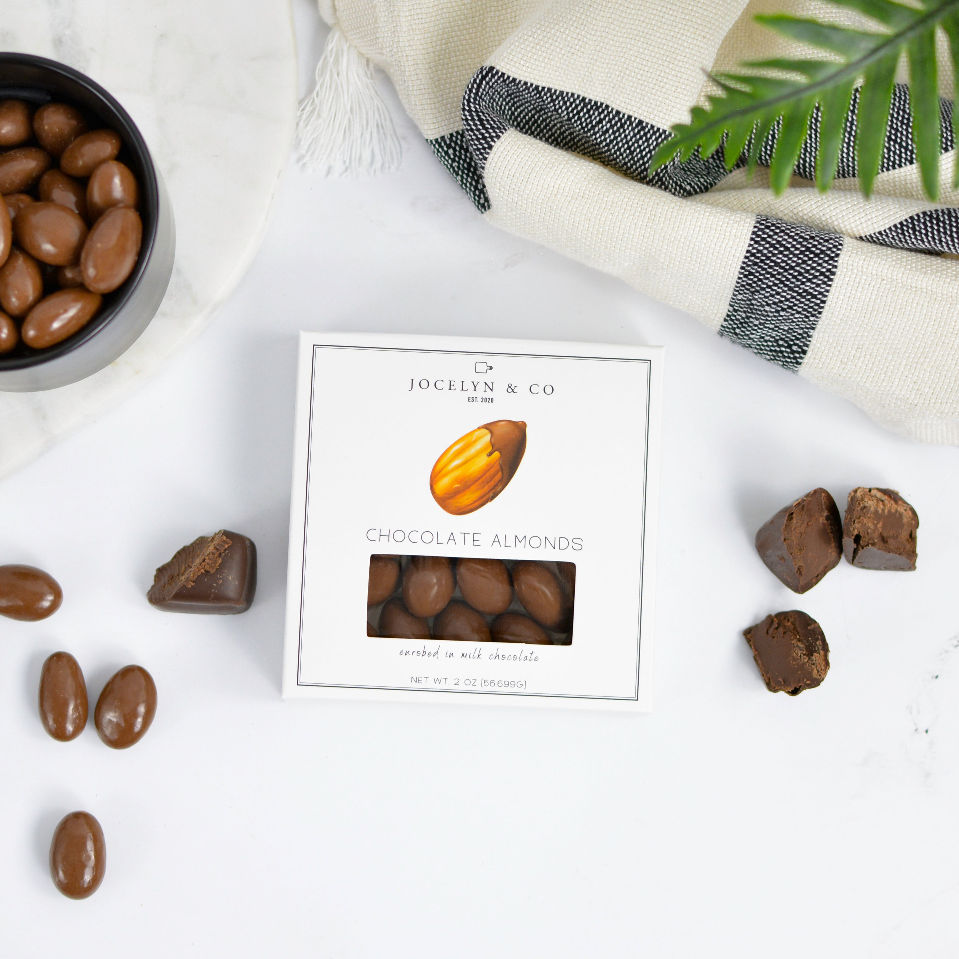 Chocolate almonds with a box on a marble surface with a striped cloth and plant in the background